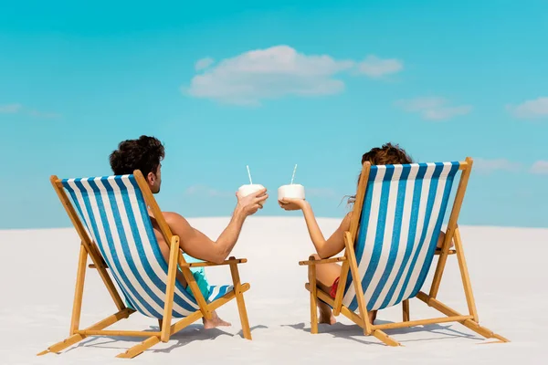 Visão traseira do jovem casal sentado em cadeiras de convés com bebidas de coco na praia de areia — Fotografia de Stock