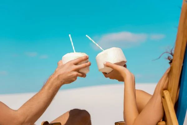 Vue recadrée du jeune couple assis dans des chaises longues avec des boissons à la noix de coco sur la plage de sable — Photo de stock