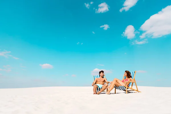 Sonriente joven pareja sentada en tumbonas en la playa de arena - foto de stock