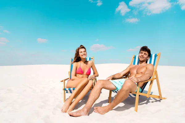 Sonriente joven pareja sentada en tumbonas en la playa de arena - foto de stock