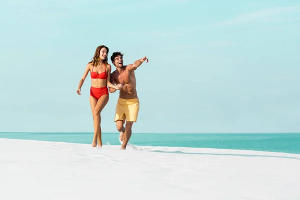 Feliz jovem casal sexy correndo na praia de areia e apontando para longe perto do oceano — Fotografia de Stock