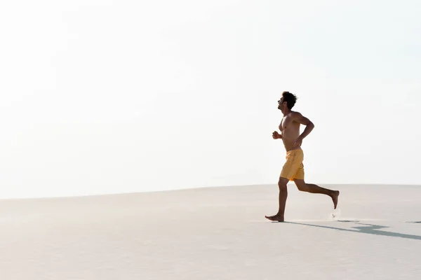 Hombre en pantalones cortos de natación con torso muscular corriendo en la playa de arena - foto de stock