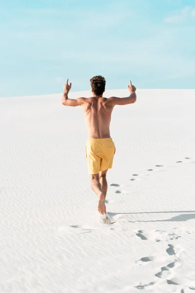 Vista trasera del hombre en pantalones cortos de natación con torso muscular corriendo en la playa de arena - foto de stock