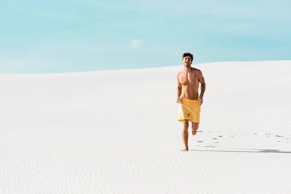 Hombre sonriente en pantalones cortos de natación con torso muscular corriendo en la playa de arena - foto de stock