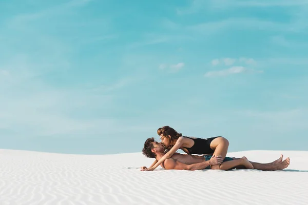 Vista lateral de la mujer joven y sexy besándose y acostado en el novio en la playa de arena - foto de stock