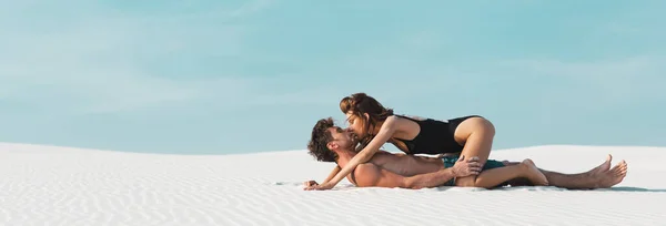 Vista lateral de la mujer joven y sexy besándose y acostado en el novio en la playa de arena, tiro panorámico - foto de stock