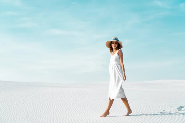 Vista lateral de la hermosa chica sonriente en vestido blanco y sombrero de paja caminando en la playa de arena con cielo azul - foto de stock