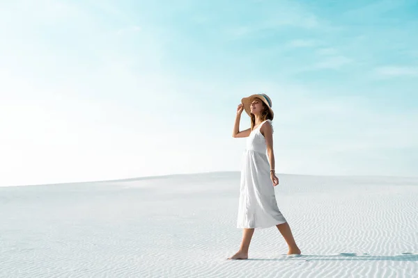 Вид сбоку улыбающейся красивой девушки в белом платье и соломенной шляпе, идущей по песчаному пляжу с голубым небом — стоковое фото