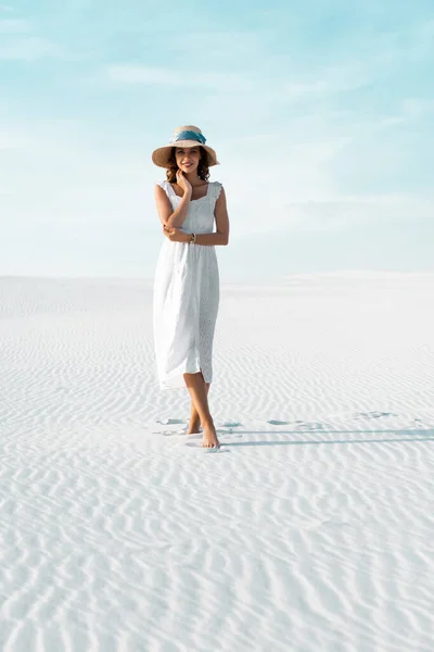 Bella ragazza in abito bianco e cappello di paglia passeggiando sulla spiaggia di sabbia con cielo blu — Foto stock