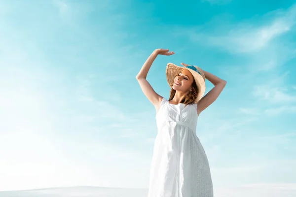 Низький кут зору усміхненої красивої дівчини в білій сукні і солом'яному капелюсі на блакитному небі — стокове фото
