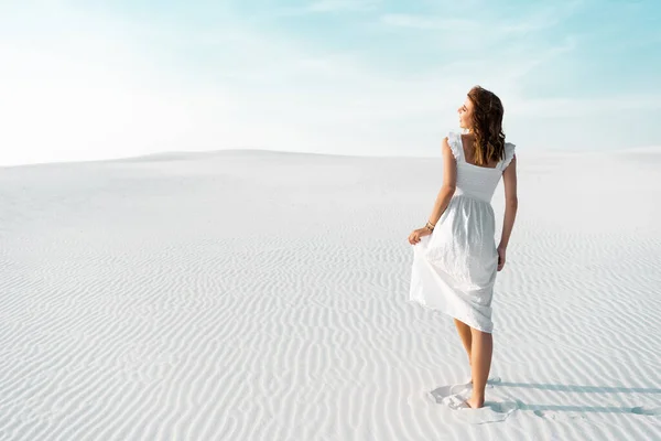 Vista trasera de la hermosa chica en vestido blanco caminando en la playa de arena con cielo azul - foto de stock