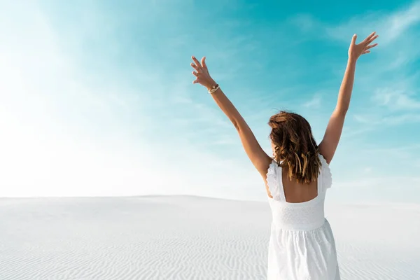 Vista trasera de hermosa chica en vestido blanco con las manos en el aire en la playa de arena con cielo azul — Stock Photo