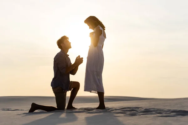 Vista lateral del joven haciendo propuesta de matrimonio a la novia en la playa de arena al atardecer - foto de stock