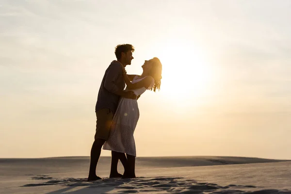 Vista lateral do jovem casal abraçando na praia de areia ao pôr do sol — Fotografia de Stock