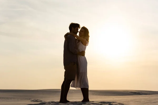 Seitenansicht eines jungen Paares, das sich bei Sonnenuntergang am Sandstrand umarmt — Stockfoto