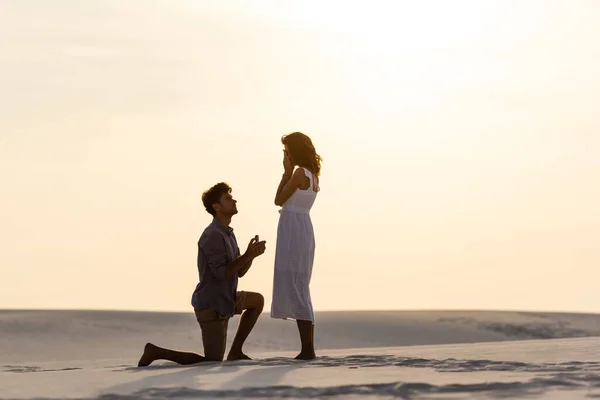 Vista lateral del joven haciendo propuesta de matrimonio a la novia en la playa de arena al atardecer - foto de stock