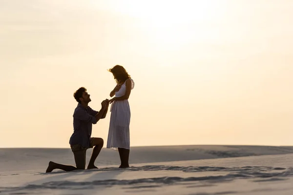 Visão lateral do jovem fazendo proposta de casamento com a namorada na praia de areia ao pôr do sol — Fotografia de Stock