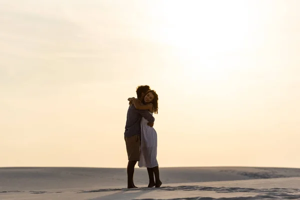 Vista lateral de pareja joven abrazándose en la playa de arena al atardecer - foto de stock