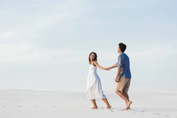 Молода пара ходить на піщаному пляжі і тримає руки на тлі блакитного неба — стокове фото