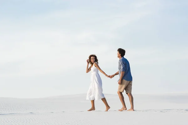 Молода пара ходить на піщаному пляжі і тримає руки на тлі блакитного неба — стокове фото