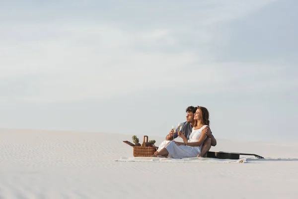 Young couple drinking champagne while sitting on blanket with basket of fruits and acoustic guitar on beach — Stock Photo