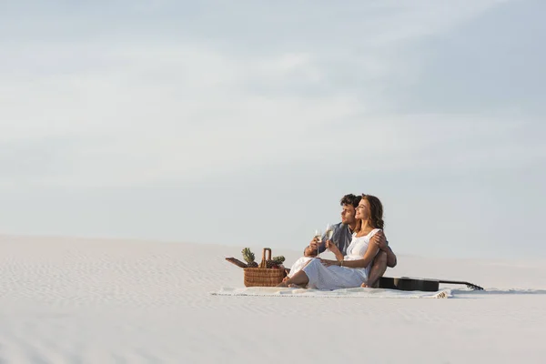 Giovane coppia bere champagne mentre seduto su una coperta con cesto di frutta e chitarra acustica sulla spiaggia — Foto stock