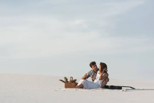 Jeune couple buvant du champagne assis sur une couverture avec panier de fruits et guitare acoustique sur la plage — Photo de stock