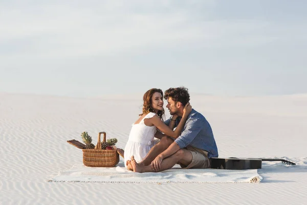 Junges Paar umarmt sich, während es auf einer Decke mit Obstkorb und Akustikgitarre am Strand sitzt — Stockfoto