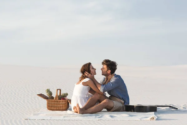Молода пара обіймається, сидячи на ковдрі з кошиком фруктів і акустичної гітари на пляжі — стокове фото