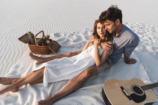 Junges Paar umarmt, während es auf einer Decke mit Obstkorb und Akustikgitarre am Strand sitzt — Stockfoto