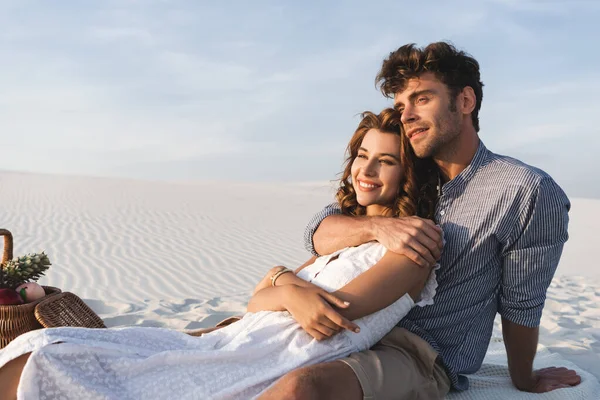 Sonriente joven pareja abrazándose mientras hace un picnic en la playa - foto de stock