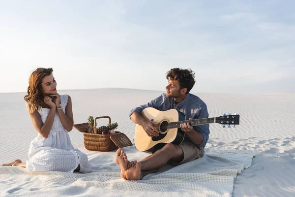 Uomo suonare la chitarra acustica alla fidanzata vicino cesto di vimini con frutta sulla spiaggia — Foto stock