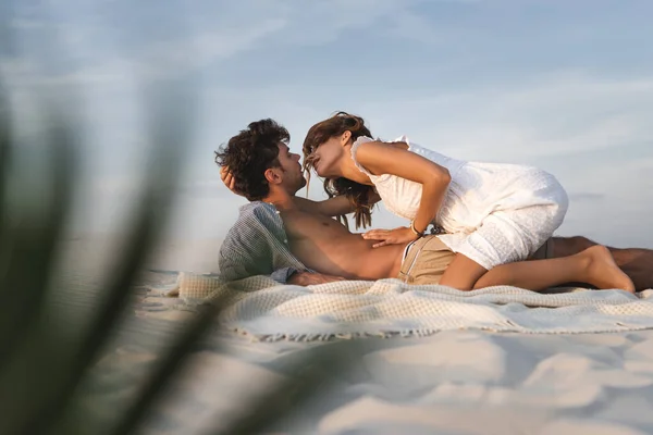 Selective focus of passionate young woman sitting on boyfriend on blanket on beach — Stock Photo