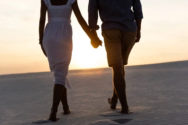 Cropped view of man and woman holding hands while walking on beach against sun during sunset — Stock Photo