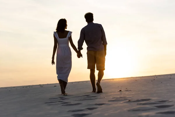 Silhouettes of man and woman holding hands while walking on beach against sun during sunset — Stock Photo