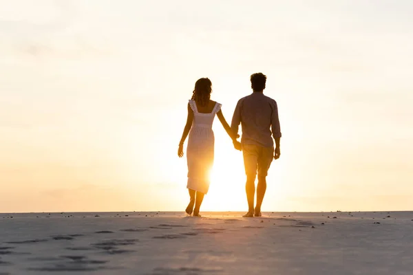 Siluetas de hombre y mujer tomados de la mano mientras caminan en la playa contra el sol durante el atardecer - foto de stock