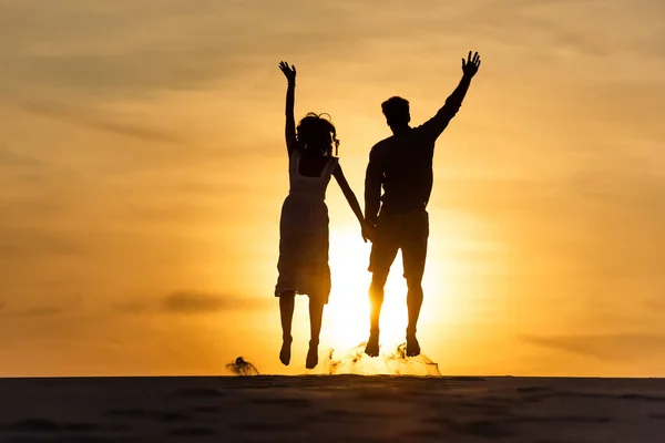 Siluetas del hombre y la mujer saltando en la playa contra el sol durante el atardecer - foto de stock