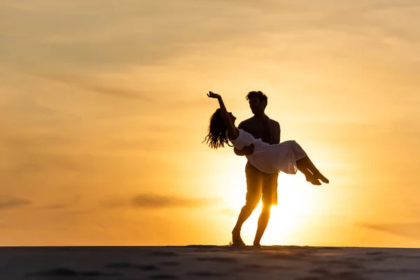 Siluetas del hombre girando alrededor de la mujer en la playa contra el sol durante la puesta del sol - foto de stock