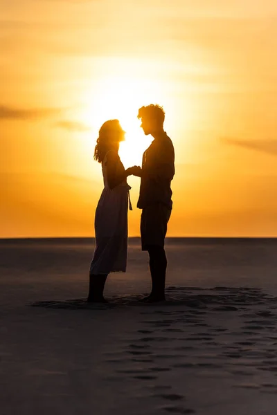 Vista lateral de las siluetas del hombre y la mujer tomados de la mano en la playa contra el sol durante la puesta del sol - foto de stock