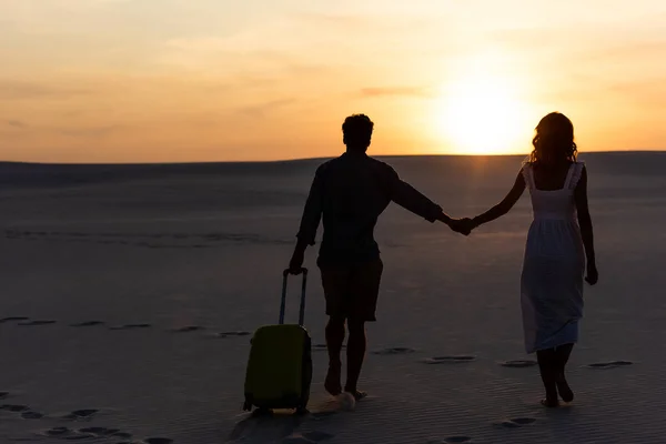 Vista trasera de la pareja caminando en la playa mientras se toma de la mano con la bolsa de viaje al atardecer - foto de stock