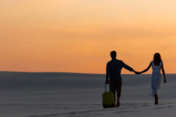 Vista trasera de la pareja caminando en la playa mientras se toma de la mano con la bolsa de viaje al atardecer - foto de stock