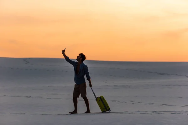 Silueta del hombre caminando en la playa con bolsa de viaje y teléfono inteligente al atardecer - foto de stock