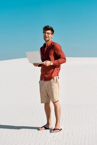 Молодой фрилансер на песчаном пляже с ноутбуком на фоне ясного голубого неба — стоковое фото