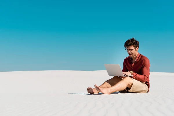 Young freelancer on sandy beach using laptop against clear blue sky — Stock Photo
