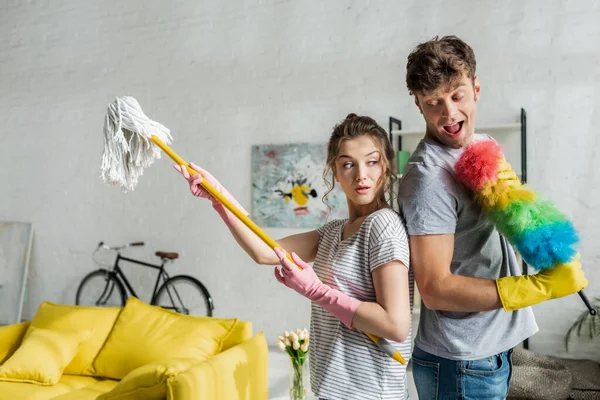 Hombre excitado y hermosa mujer sosteniendo fregona y cepillo de plumero en casa — Stock Photo