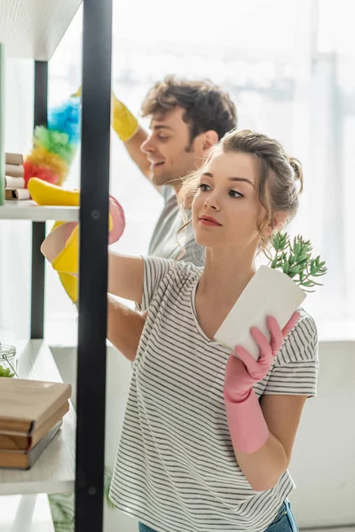 Selective focus of attractive girl in rubber gloves holding plant while cleaning shelve with rag near man — Stock Photo