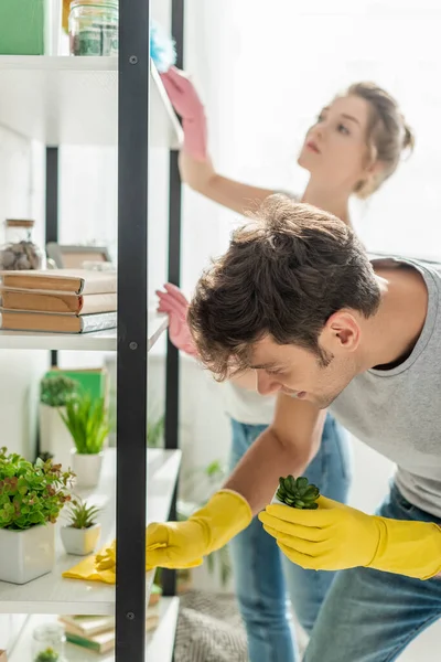 Selective focus of handsome man holding plant while cleaning shelve with rag near woman — Stock Photo