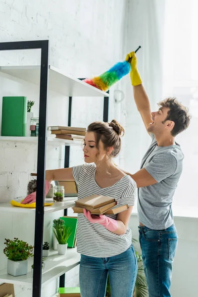 Attraktives Mädchen in Gummihandschuhen, das Bücher hält, während es Regale mit Lappen in der Nähe eines Mannes säubert — Stockfoto