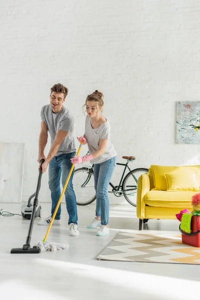 Hombre feliz usando aspiradora cerca de mujer atractiva lavando piso con fregona - foto de stock