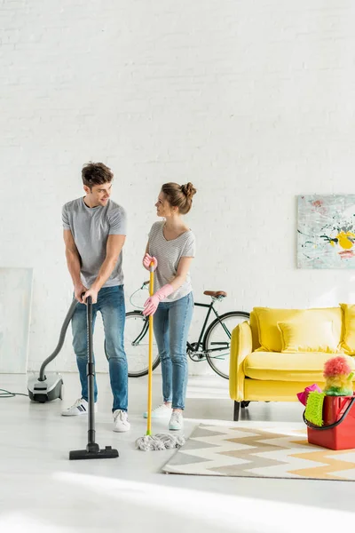 Happy man using vacuum cleaner near woman washing floor with mop — Stock Photo